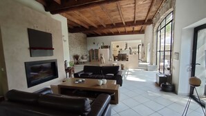 A spacious living area with a high ceiling, a fireplace, a television, and a view into the kitchen.