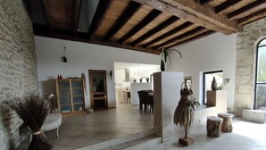 A spacious room with a high ceiling, stone walls, and a tiled floor. There is a wooden door, a dining area with chairs and a table, and a decorative plant.