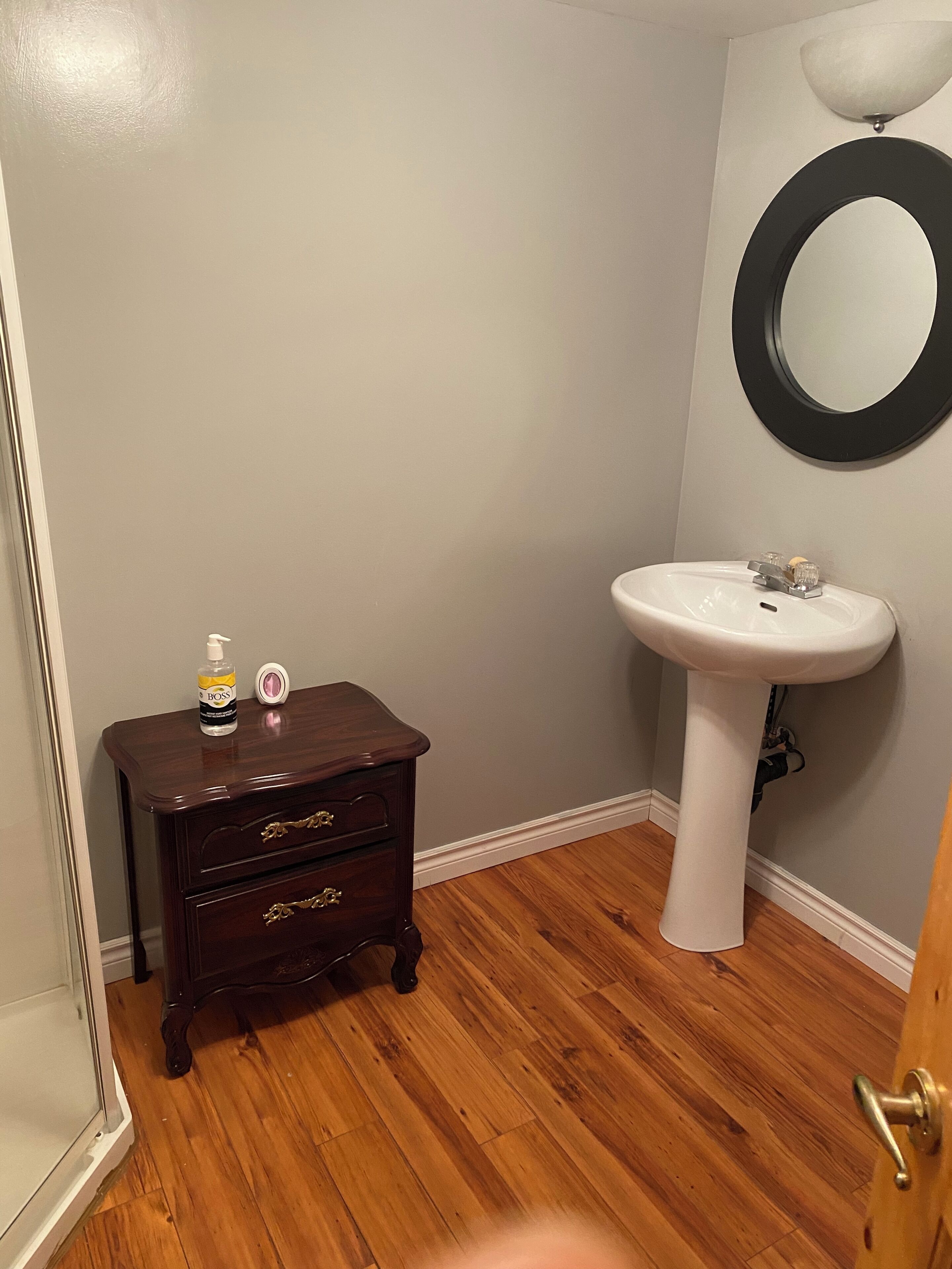 A bathroom with a wooden floor, a white sink, a dark wooden dresser, and a round mirror on the wall.