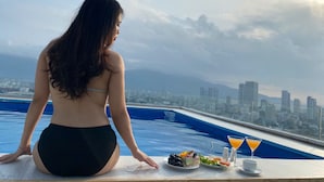 Une personne assise au bord d’une piscine, avec une vue sur la ville, une table avec de la nourriture et des boissons, et un ciel dégagé.