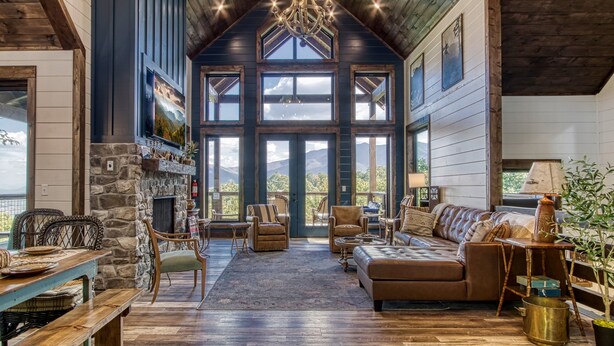 A spacious living room with a stone fireplace, large windows, and a high ceiling with exposed beams.