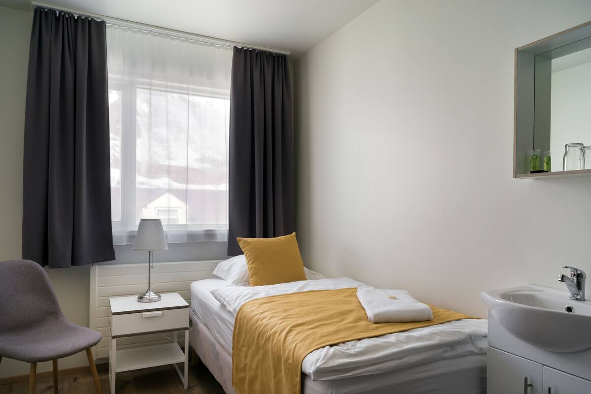 A hotel room with a bed, a bedside table with a lamp, a chair, a window with curtains, and a bathroom with a sink and mirror.