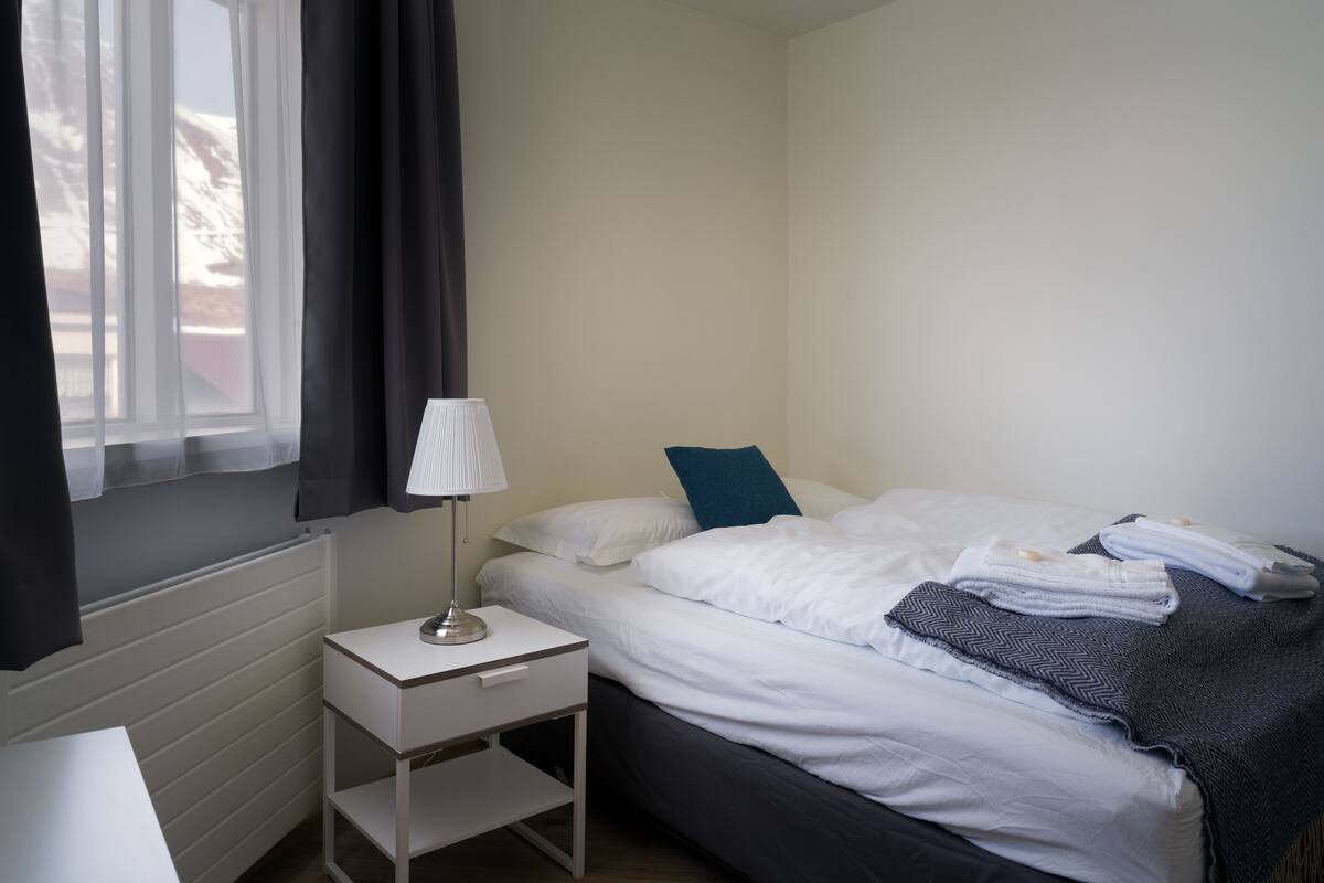 A bedroom with a bed, a bedside table with a lamp, a window with curtains, and a radiator.