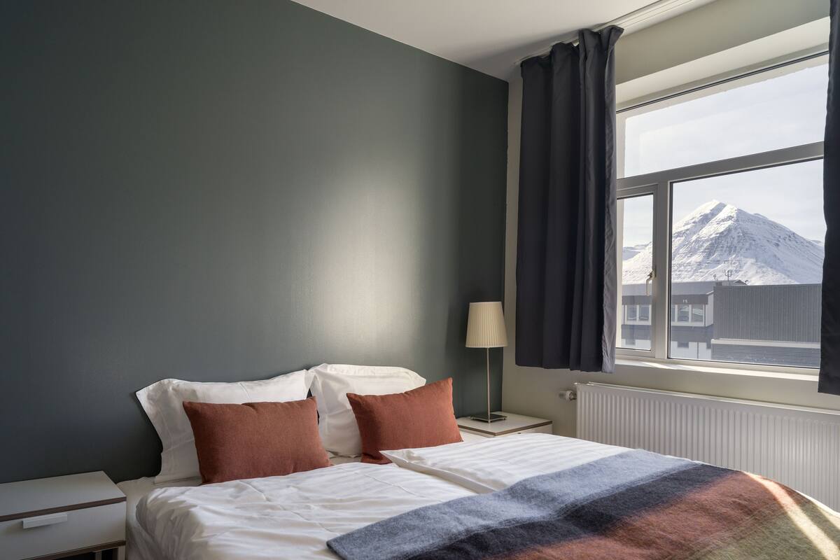 A hotel room with a bed, a nightstand, a lamp, and a window with a view of a mountain.