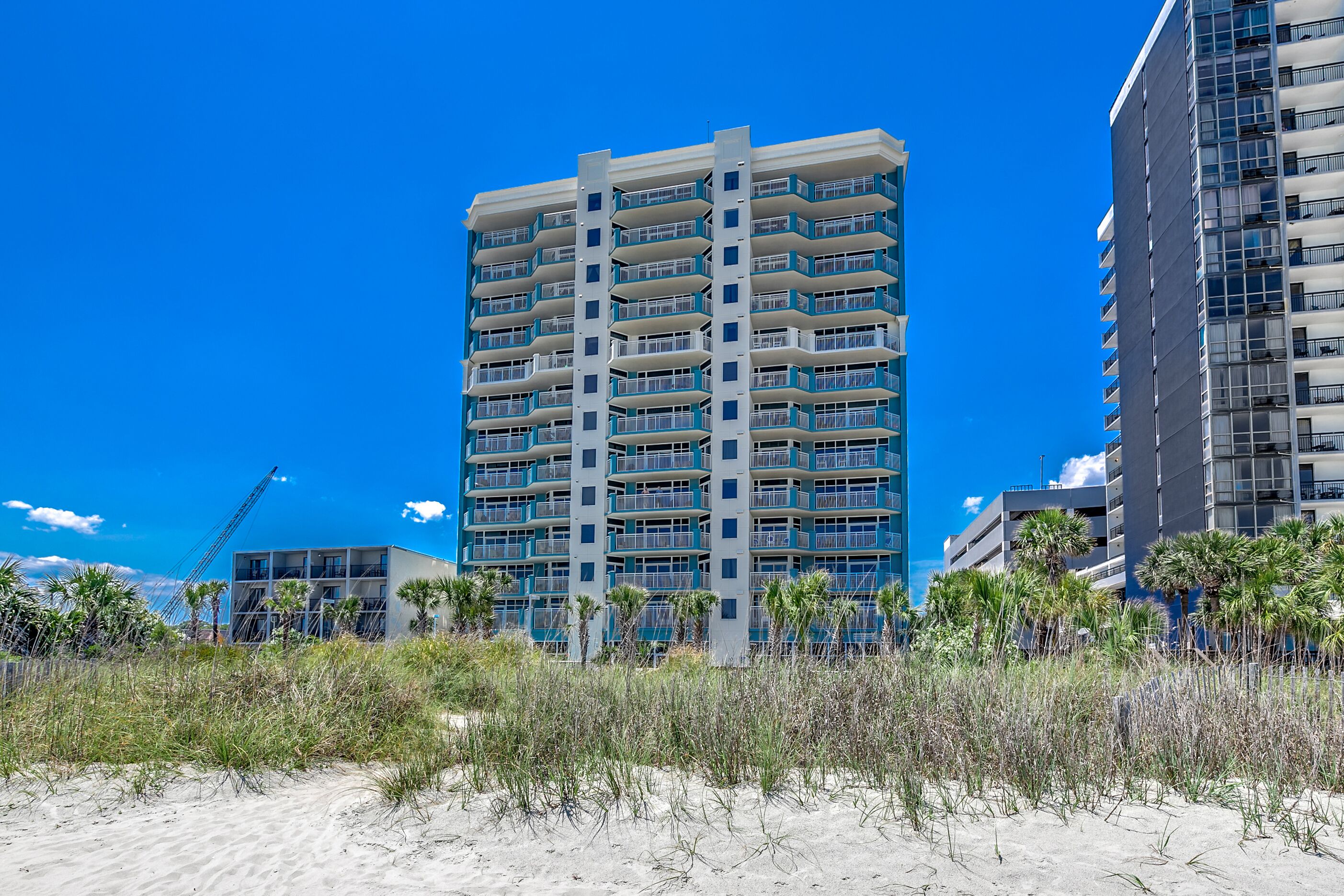 A high-rise hotel with multiple balconies, located near a beach with sand and sparse vegetation.