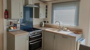 A compact kitchen with a gas stove, oven, and a window with blinds. - The Sea Breeze - Captivating Caravan in Camber (Rye)