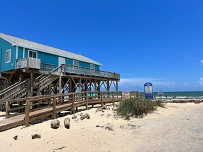 A beach house with a wooden deck and stairs leading to a balcony, a sign indicating a beach access point, and a clear view of the ocean. - Sea Forever - Front Row, Fire Pit & Miles of Surf! (Surfside Beach)