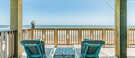 A wooden deck with two chairs and a patterned ottoman, overlooking a beach.