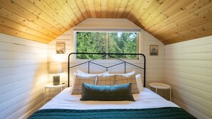 Un dormitorio con techo inclinado, una cama con varias almohadas, una mesita de noche con una lámpara y una ventana con vistas a árboles.