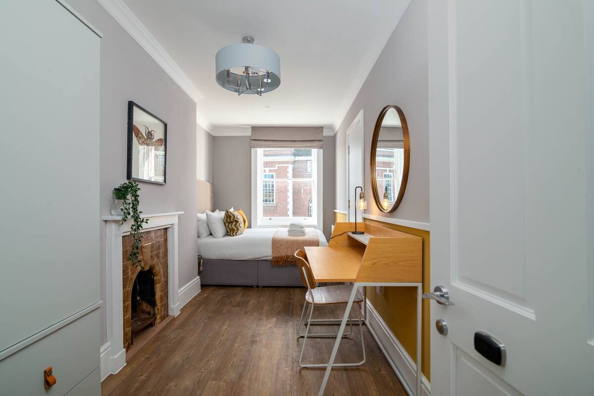 A bedroom with a bed, a desk, a chair, a fireplace, a window, and a door.