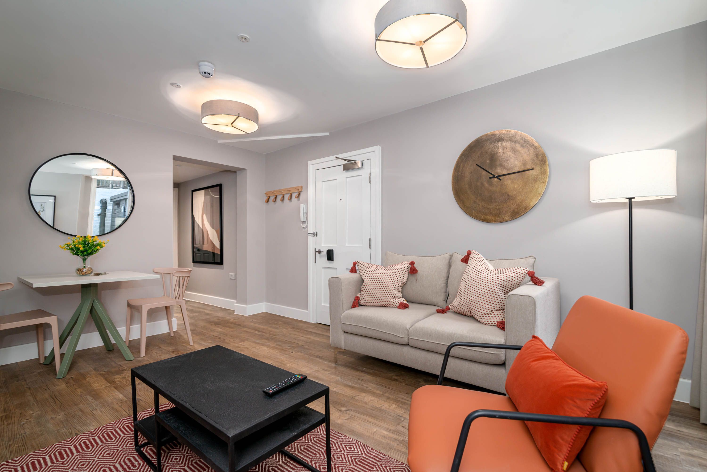 A modern living room with a sofa, a coffee table, a dining table with chairs, a large mirror, and a clock on the wall.