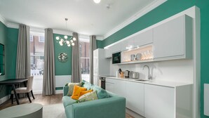 A modern living room with a teal sofa, a dining area with a table and chairs, and a kitchen with white cabinets and a built-in oven. - 37 Doughty Street (London)