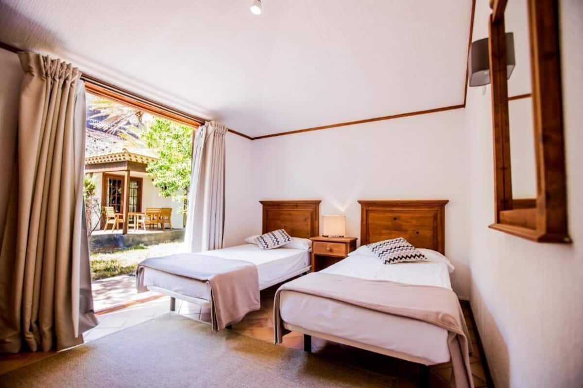 Una habitación de hotel con dos camas individuales, un cabecero de madera, una mesita de noche, una ventana con cortinas y vistas a un patio exterior.