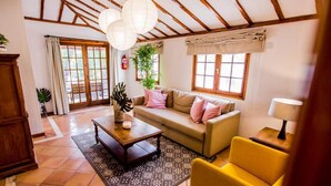 Una sala de estar acogedora con sofá, sillón amarillo y una mesita de centro. Hay dos ventanas con cortinas, una planta y un techo de madera.
