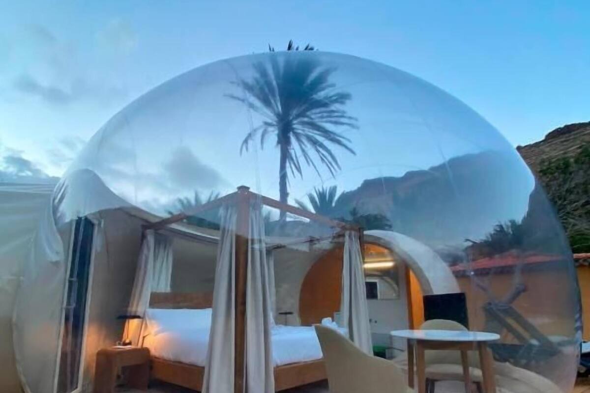 Una cúpula transparente que alberga una cama, una silla y una mesa, reflejando una palmera y montañas en el exterior.