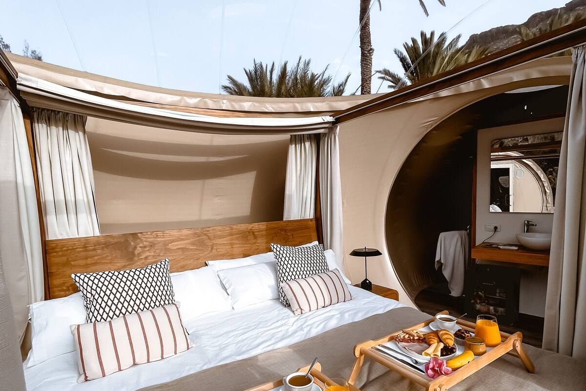 Un dormitorio con cama, almohadas, una bandeja con alimentos para el desayuno y vistas a palmeras.