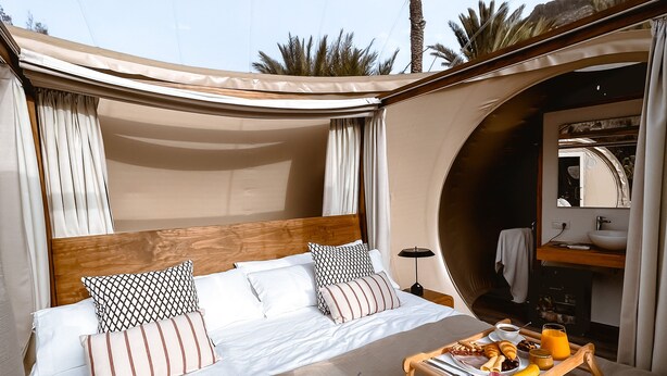 Un dormitorio con cama, almohadas, una mesita de noche con una bandeja de desayuno y vistas a palmeras.