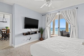 A hotel room with a bed, a TV mounted on the wall, a dining area with a table and chairs, and a balcony with curtains.