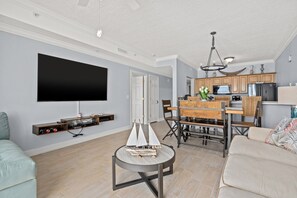 A modern living room with a flat-screen TV, a round coffee table with a model sailboat, a sofa, and a dining area with a wooden table and chairs.