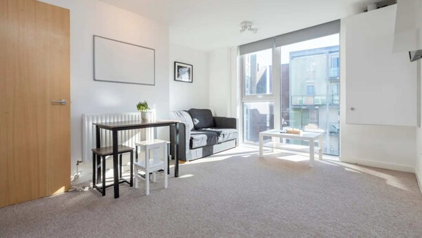 Una sala de estar moderna con un sofá gris, una mesa de centro blanca y una pequeña mesa con sillas. Hay un radiador, un cuadro enmarcado en la pared y una ventana con vistas a edificios.