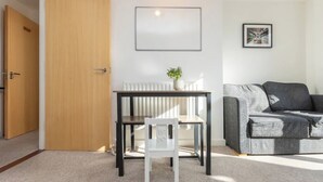 Una habitación con puerta de madera, una silla blanca, una mesa con una planta, un sofá gris y un cuadro en la pared.