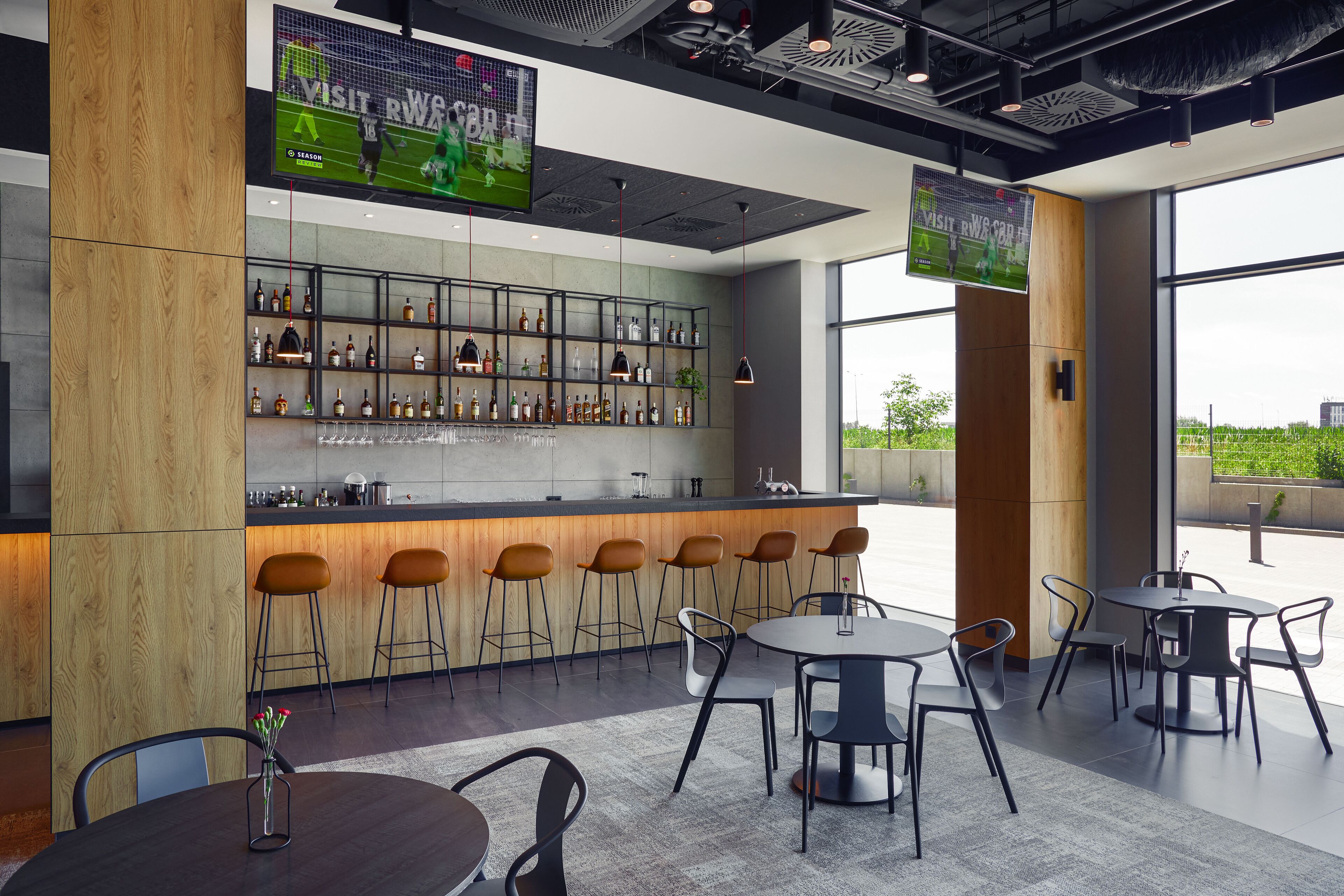 A modern bar area with a wooden bar counter, multiple stools, and a large TV screen displaying sports.