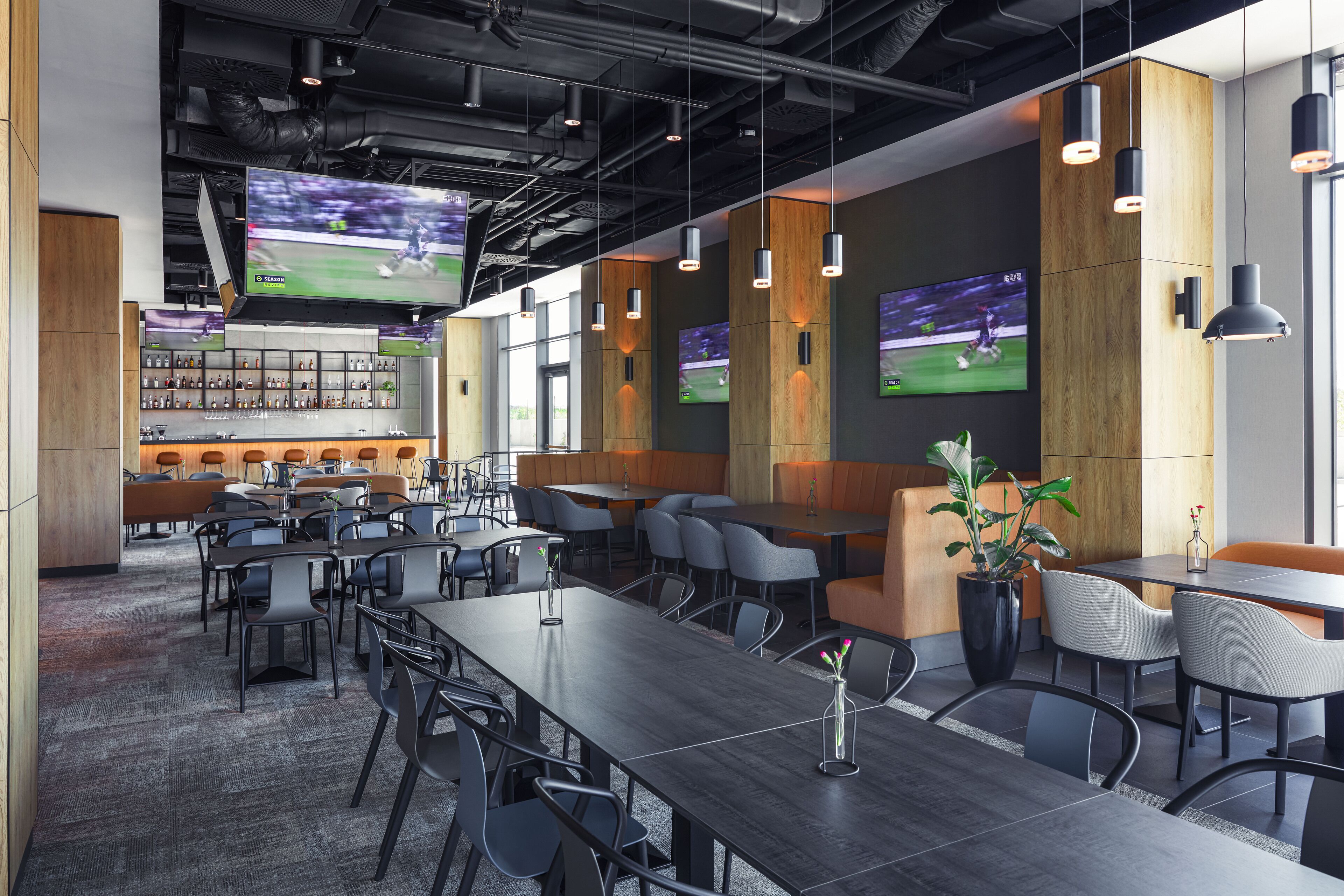 A modern conference room with multiple tables and chairs, a bar area with shelves of bottles, and large screens displaying sports events.
