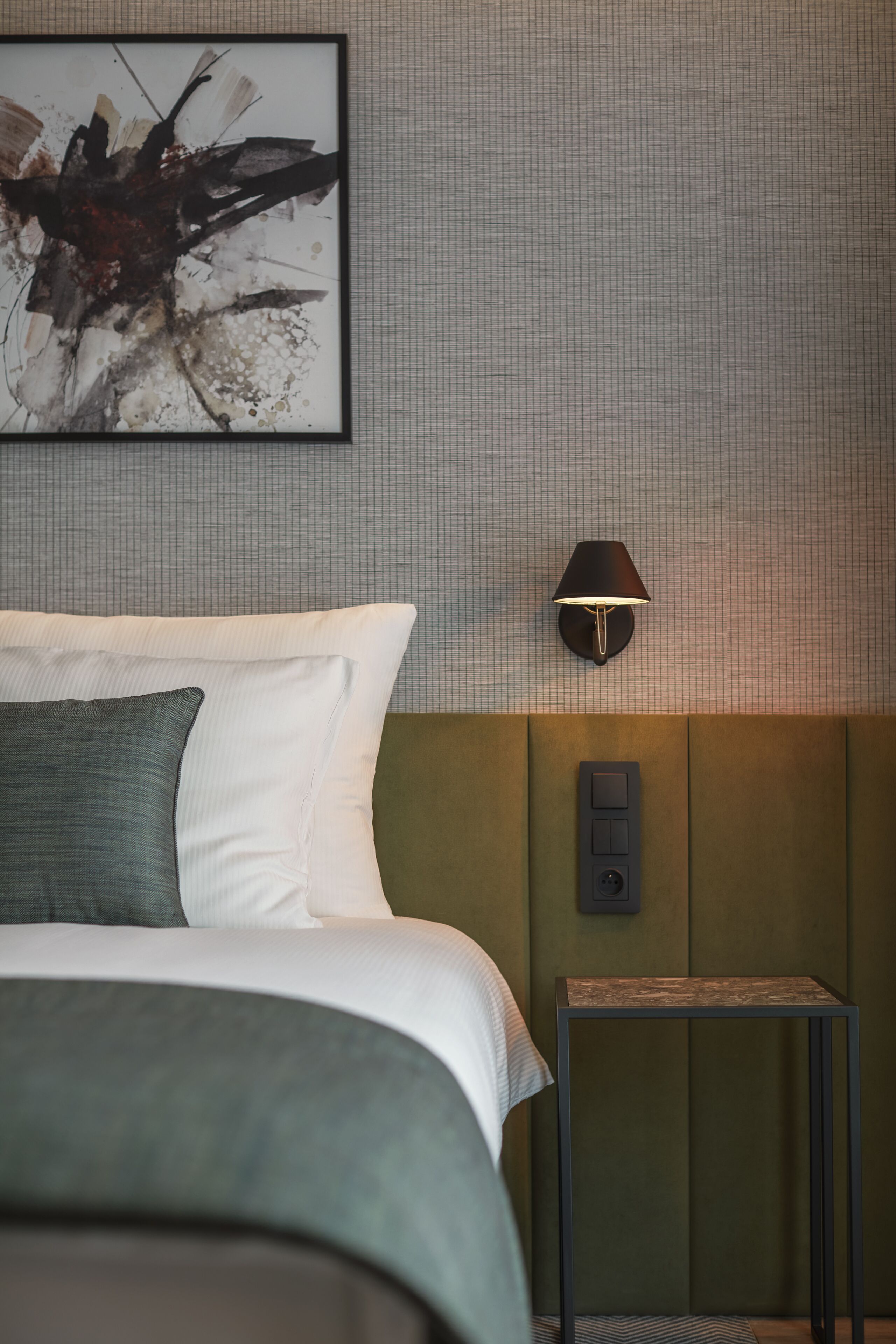A neatly made bed with pillows, a wall-mounted lamp, and a small table beside it.