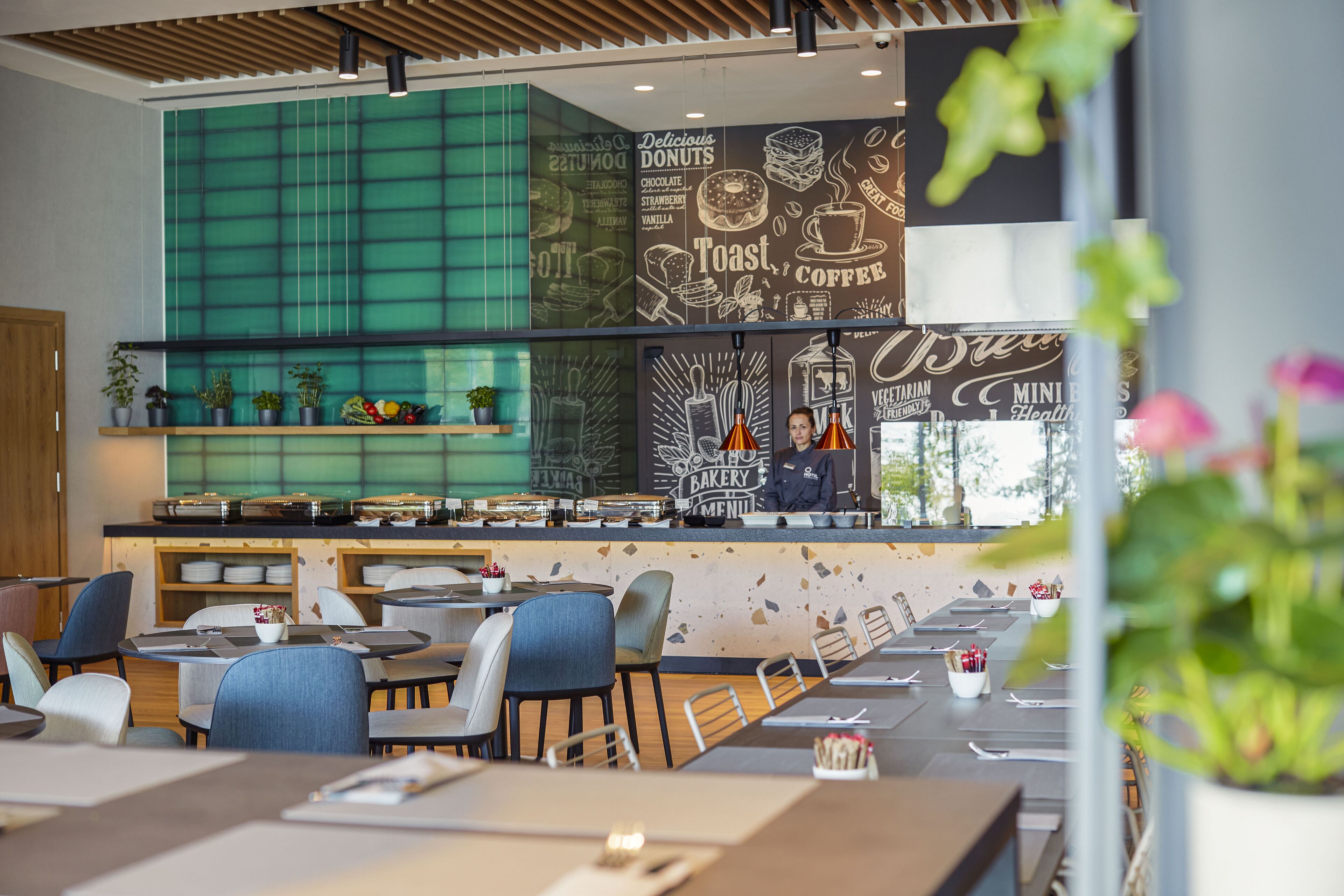 A modern cafe with a green-tiled wall, a bakery menu, and a coffee menu. There are tables and chairs arranged for dining.