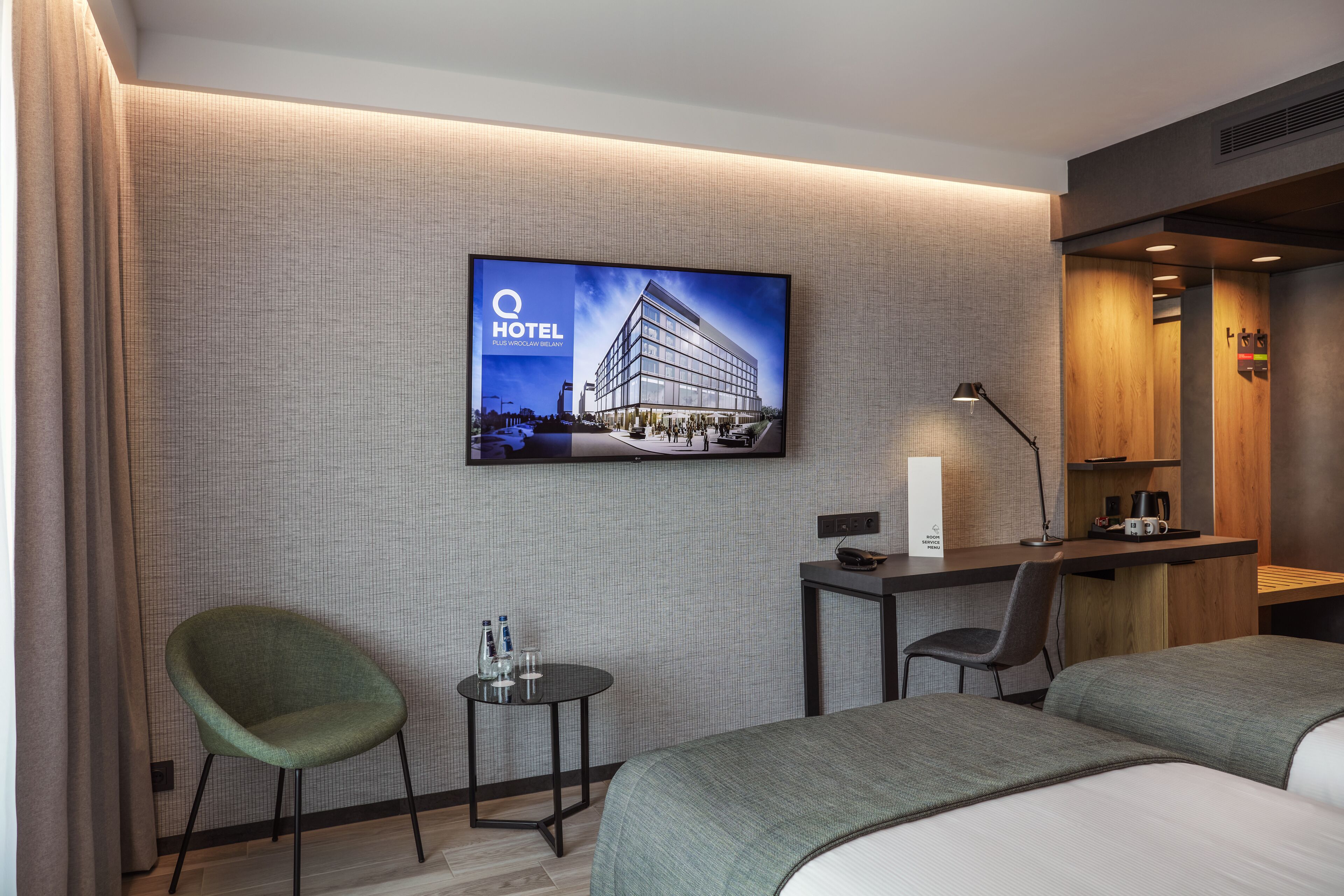 Une chambre d’hôtel avec un lit, un bureau, une chaise, un téléviseur fixé au mur et une petite table avec une lampe.