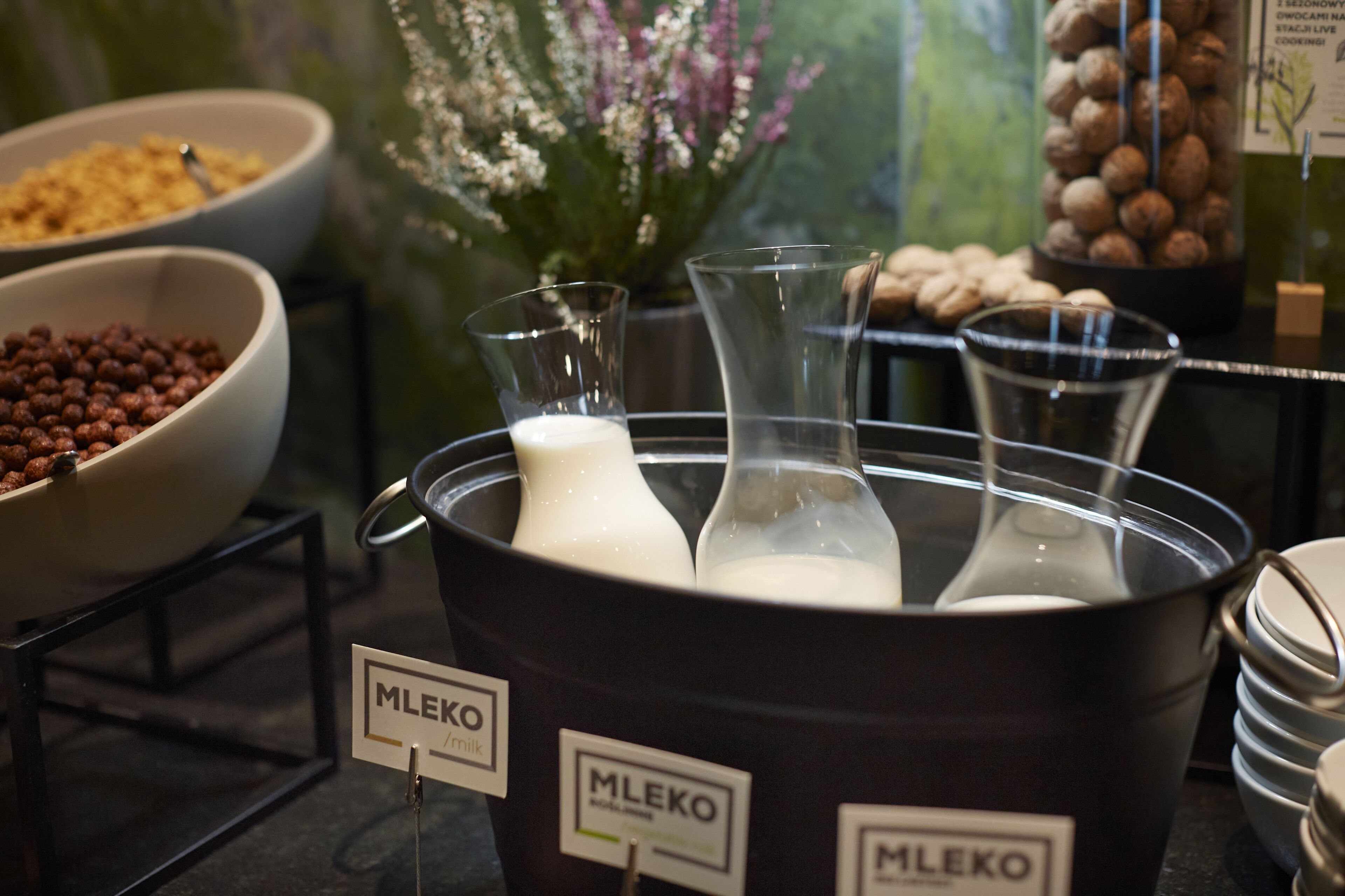 A buffet setup with labeled containers of MLEKO milk, a bowl of cereal, and a vase of flowers.