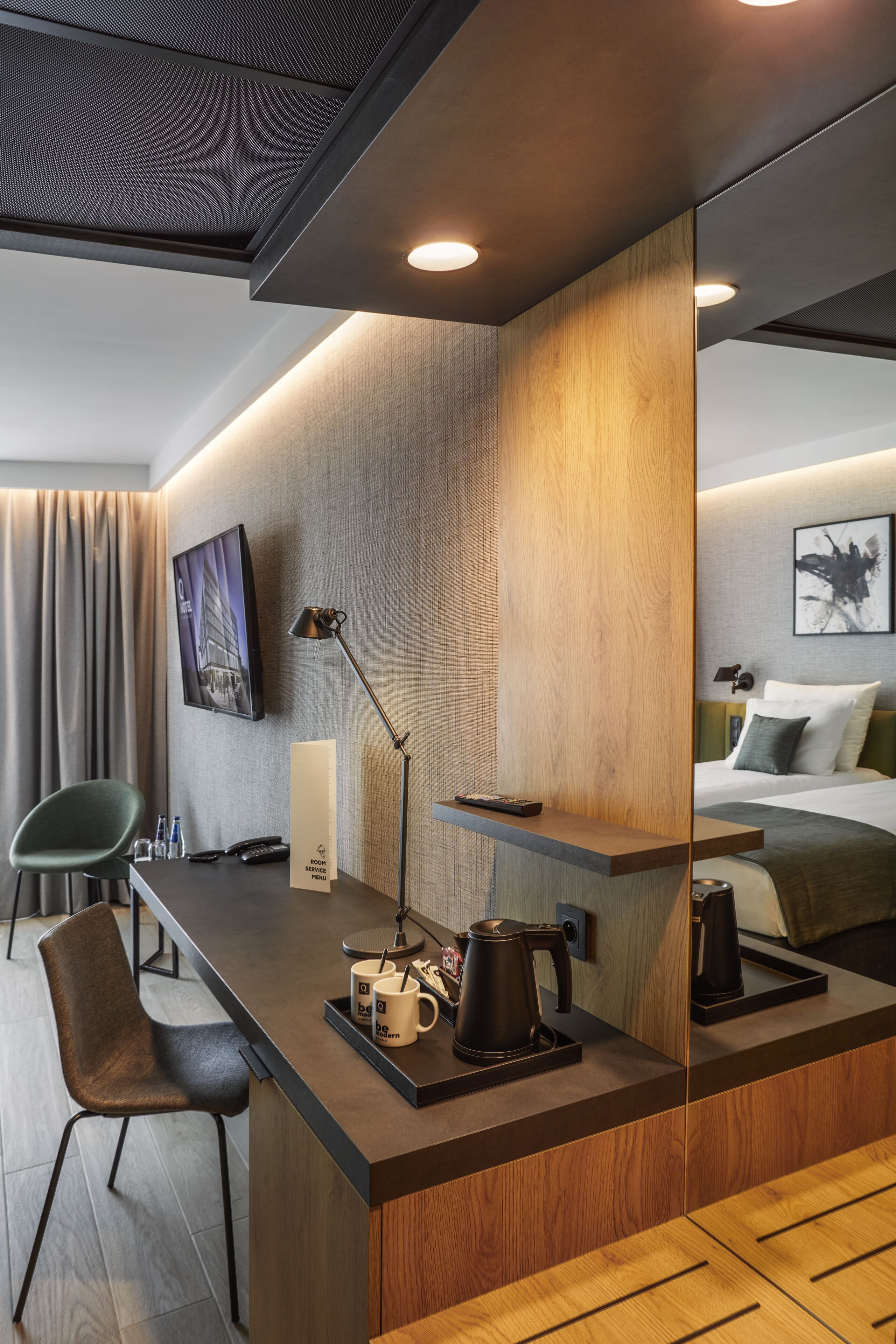 A modern hotel room with a wooden desk, a chair, a bed, a bedside table, a lamp, and a wall-mounted TV.