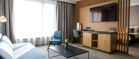 Moderni hotellihuone, jossa on sohva, sohvapöytä, työpöytä ja tasatelevisio sekä sänky.