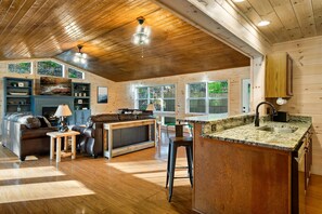 Amplia sala de estar con techo de madera, chimenea y cocina con encimera de granito y taburetes de bar.