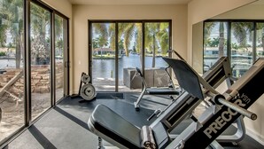 Une salle de sport équipée d’un tapis de course, d’un vélo d’appartement et de poids. Elle possède de grandes fenêtres offrant une vue sur des palmiers et une piscine.