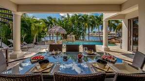 Une table avec un plateau en verre, dressée pour le repas, donnant sur la piscine, entourée de chaises en osier et surmontée d’un parasol en chaume.