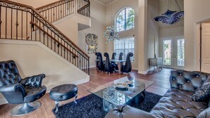 Un vaste salon comprenant un canapé en cuir noir, une table basse en verre et un fauteuil en cuir noir. Un escalier orné d’une rampe en fer forgé mène à l’étage supérieur.