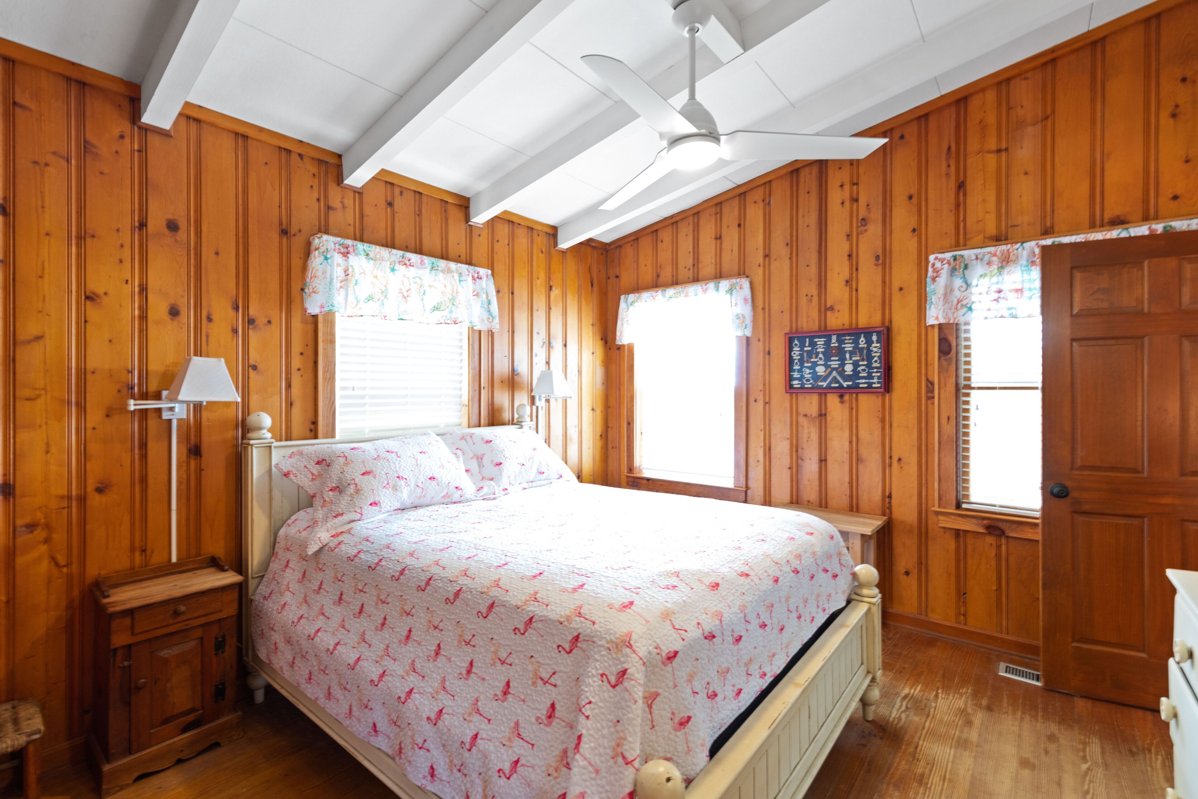 Dormitorio con paneles de madera, una cama, una mesita de noche, una lámpara y un ventilador de techo.