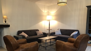 Un salon avec deux canapés, une table basse et deux fauteuils. On aperçoit une lampe sur une table d’appoint et une bibliothèque au fond de la pièce.