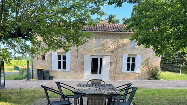 Un bâtiment rustique en pierre, avec un toit en tuiles, une terrasse en bois dotée d’une table et de chaises, et une pelouse à l’avant.