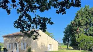 Un bâtiment rustique en pierre, doté d’un toit en tuiles, d’un petit porche et d’une cour verdoyante aménagée avec des sièges extérieurs.