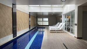 A modern indoor swimming pool with clear blue water, a wooden floor, and white sun loungers along the side.