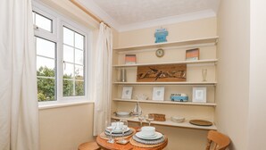 A dining area with a round wooden table, two chairs, and a window with curtains. Shelves above the table hold various decorative items and a clock. - 16 Norfolk Heights (King's Lynn)
