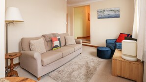 A living room with a beige sofa, a blue armchair, a wooden side table, a lamp, and a painting on the wall. - 16 Norfolk Heights (King's Lynn)