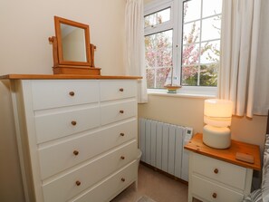 A bedroom with a white dresser, a small bedside table with a lamp, a window with pink flowers outside, and a radiator. - 16 Norfolk Heights (King's Lynn)