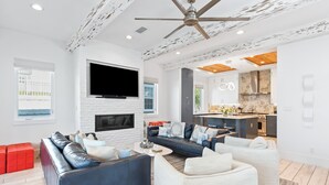A modern living room with a fireplace, a large TV, and a kitchen area visible through an open door. - Gulf Views, LSV, Pool and 2 Blocks to Beach (Santa Rosa Beach)