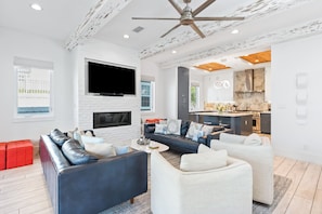 A modern living room with a fireplace, a large TV, and a kitchen area visible through an open door. - Luxury Seagrove Home with Private Pool, Rooftop Spa, Elevator, and LSV (Santa Rosa Beach)