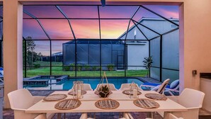 Uma área de refeições com vista para uma piscina e uma casa, através de uma estrutura de janela geométrica.