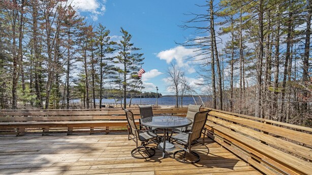 A wooden deck with a round table and chairs, overlooking a body of water and surrounded by trees.