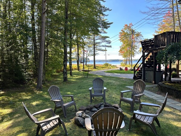 Un patio con sillas Adirondack, una fogata y vistas a un lago.